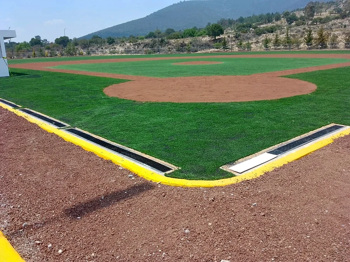 Esquina del campo de béisbol con pasto sintético, guarnición amarilla y canaleta de drenaje en Santiago Tenango, Puebla