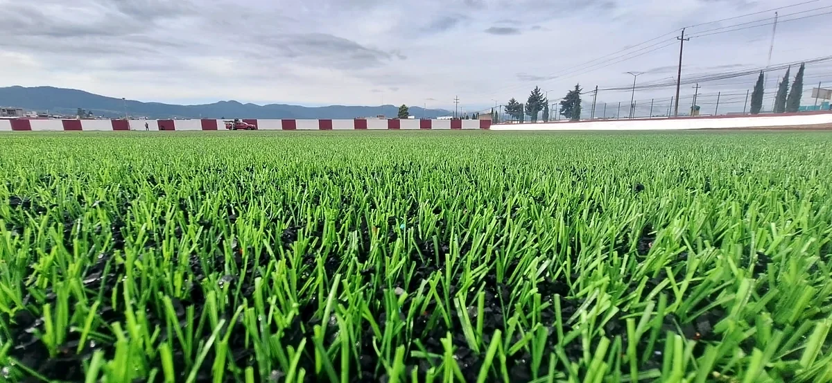 Pasto sintético a nivel de piso en la cancha de soccer del Campo Deportivo Chignahuapan, Puebla, con postes de iluminación al fondo