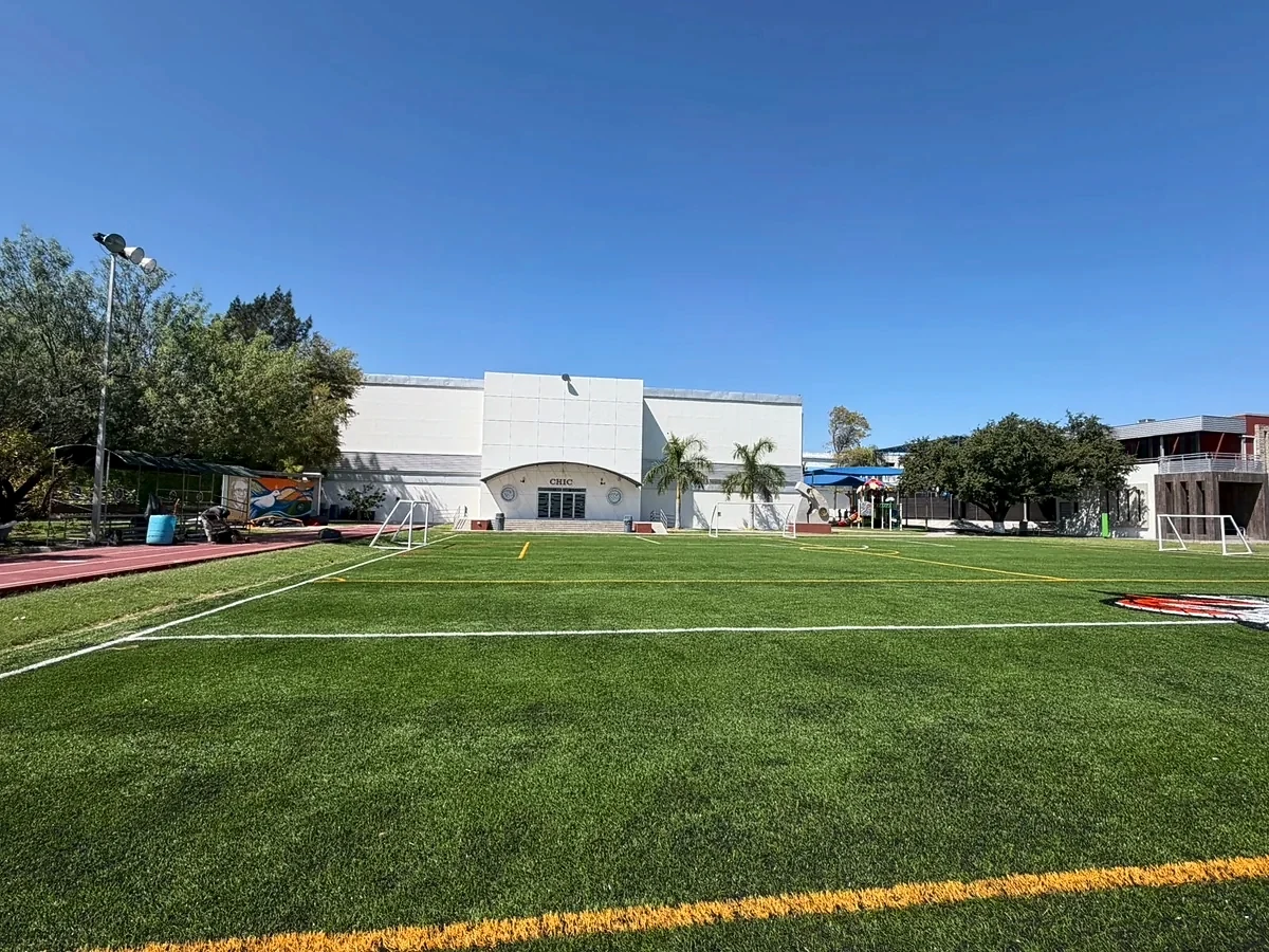 Cancha de fútbol 7 con pasto sintético del Colegio Mexicano en Reynosa, con el edificio del colegio y poste de iluminación al fondo