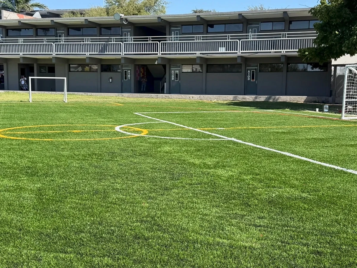 Cancha de pasto sintético con líneas blancas y amarillas frente a los salones de un colegio en Reynosa